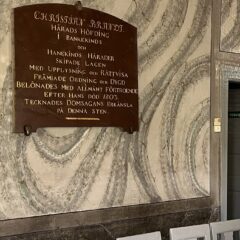 En mönstrad stenvägg med en mörk minnestavla i trä ovanför en rad ljusgrå stolar i ett äldre rum.