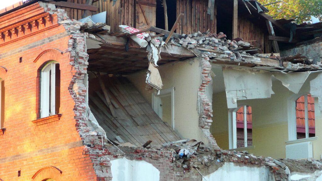 En kraftigt raserad tegelbyggnad där en hel fasad har kollapsat och inre rum med takbjälkar och väggrester ligger öppet mot utsidan.