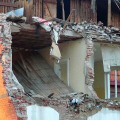 En kraftigt raserad tegelbyggnad där en hel fasad har kollapsat och inre rum med takbjälkar och väggrester ligger öppet mot utsidan.