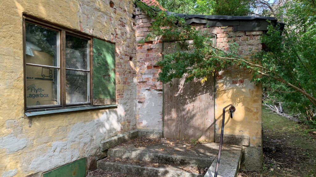 En solbelyst, förfallen husfasad i gul puts med trasiga tegelpartier, där en igensatt dörr och en övervuxen buske skymmer entrén.