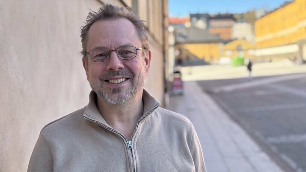 Leende man med glasögon och grå tröja står utomhus.