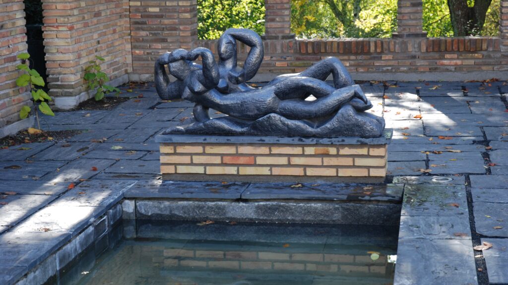 Abstrakt mörk skulptur av två sammanflätade figurer på en låg tegelsockel intill ett grunt vattenkar på en stenlagd uteplats.