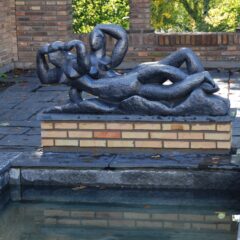 Abstrakt mörk skulptur av två sammanflätade figurer på en låg tegelsockel intill ett grunt vattenkar på en stenlagd uteplats.