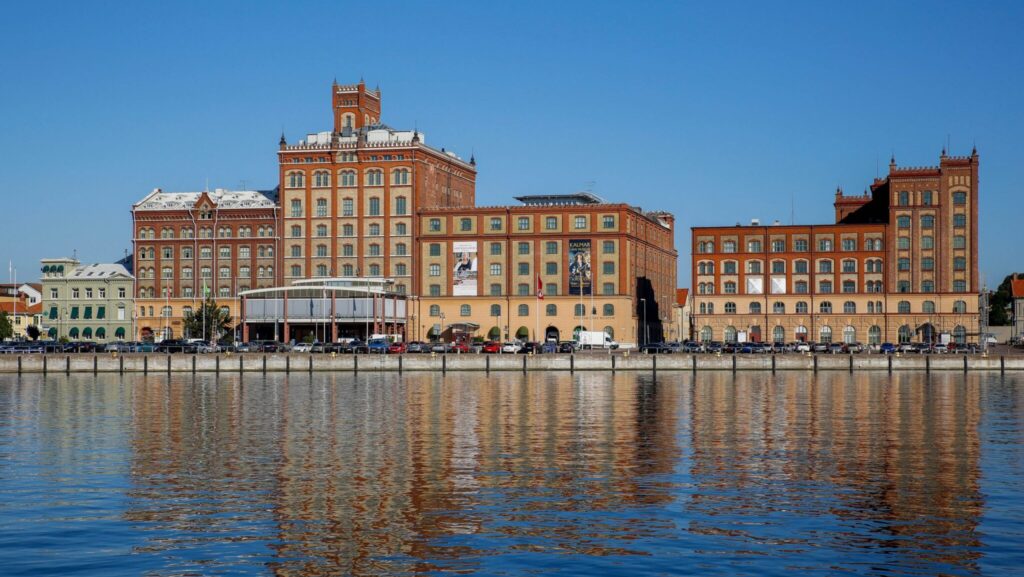 Kalmar läns museum och intilliggande historiska tegelbyggnader vid lugnt vatten med spegelbild under klarblå himmel.