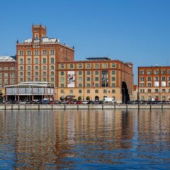 Kalmar läns museum och intilliggande historiska tegelbyggnader vid lugnt vatten med spegelbild under klarblå himmel.