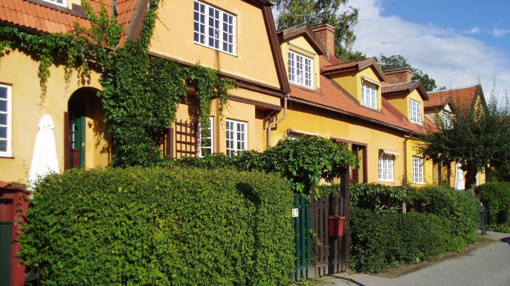 Gula radhus med vita fönster och röda tegeltak längs en solig gata, omgivna av gröna häckar och klängväxter.