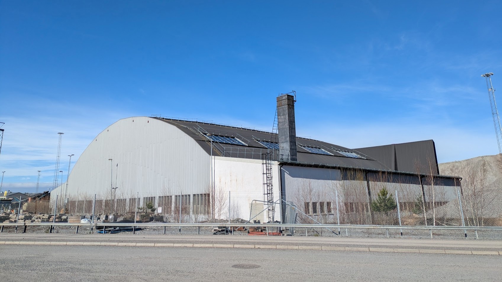 Stor bågformad industribyggnad i ljus plåt med hög skorsten och glaspartier på taket, sedd från en väg under klar blå himmel.