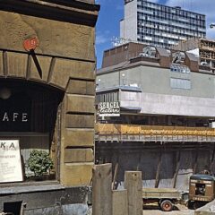 En gulbrun äldre husfasad med ett kaféfönster i förgrunden och en stor byggarbetsplats med höga ställningar och kranar i bakgrunden.