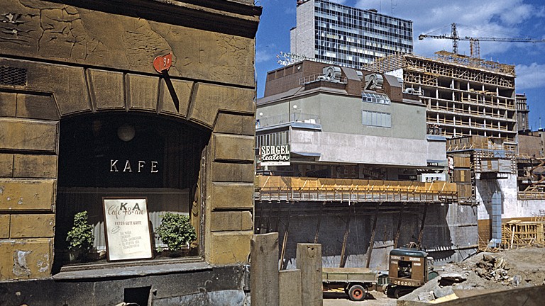 En gulbrun äldre husfasad med ett kaféfönster i förgrunden och en stor byggarbetsplats med höga ställningar och kranar i bakgrunden.