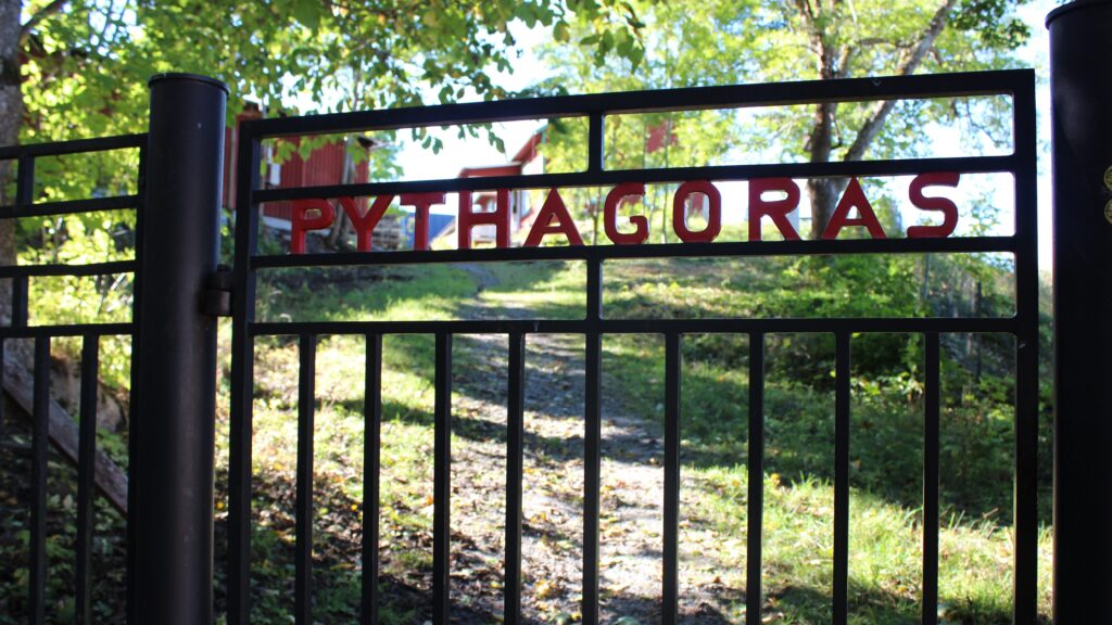 Svart metallgrind med röda bokstäver som bildar ordet Pythagoras och en solbelyst grusgång upp mot röda byggnader bakom.