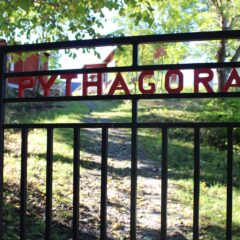 Svart metallgrind med röda bokstäver som bildar ordet Pythagoras och en solbelyst grusgång upp mot röda byggnader bakom.