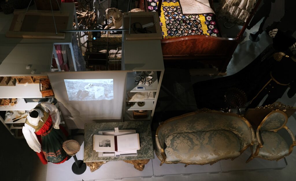 Ett museimagasin med en blandad samling föremål, bland annat två skyltdockor i historiska dräkter, äldre möbler, en träbädd med virkat täcke, hyllor med kulturföremål och en svartvit projektion på väggen.