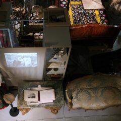 Ett museimagasin med en blandad samling föremål, bland annat två skyltdockor i historiska dräkter, äldre möbler, en träbädd med virkat täcke, hyllor med kulturföremål och en svartvit projektion på väggen.