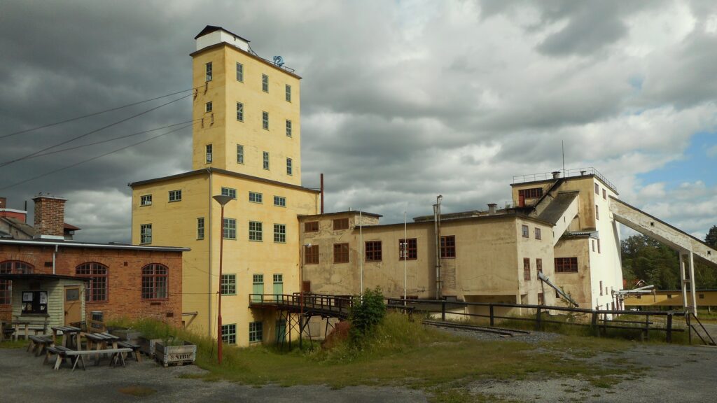 Gul och beige industribyggnad med högt torn, lägre tegelbyggnader och transportband under molnig himmel i ett öppet gruvområde.