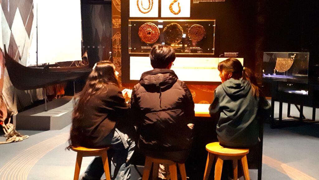 Tre skolelever sitter på pallar och tittar på saker i en utställningshall.