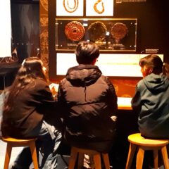 Tre skolelever sitter på pallar och tittar på saker i en utställningshall.