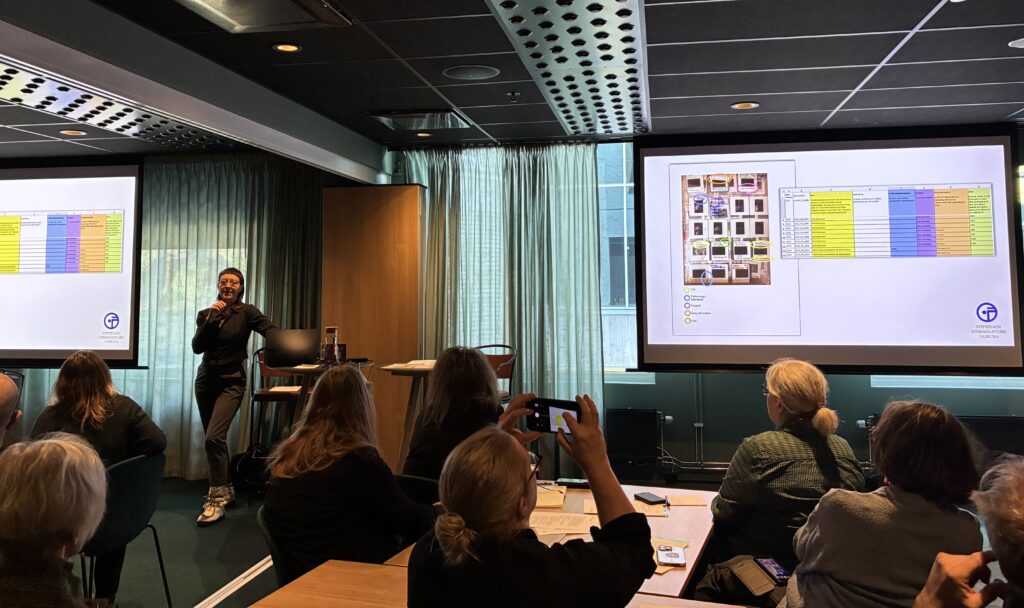 En person presenterar något på en skärm framför en grupp människor i en lokal.