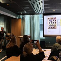 En person presenterar något på en skärm framför en grupp människor i en lokal.