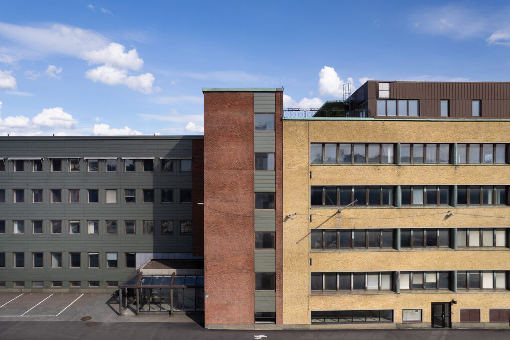 Fasad av en fler­våningsbyggnad med tre sammanhängande delar i grön, röd och gul tegel, med rader av fönster och blå himmel ovanför.