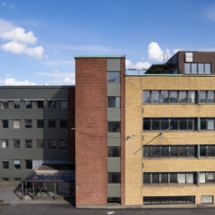 Fasad av en fler­våningsbyggnad med tre sammanhängande delar i grön, röd och gul tegel, med rader av fönster och blå himmel ovanför.