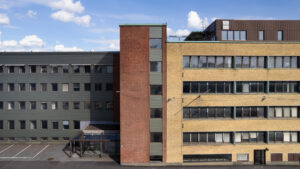 Fasad av en fler­våningsbyggnad med tre sammanhängande delar i grön, röd och gul tegel, med rader av fönster och blå himmel ovanför.