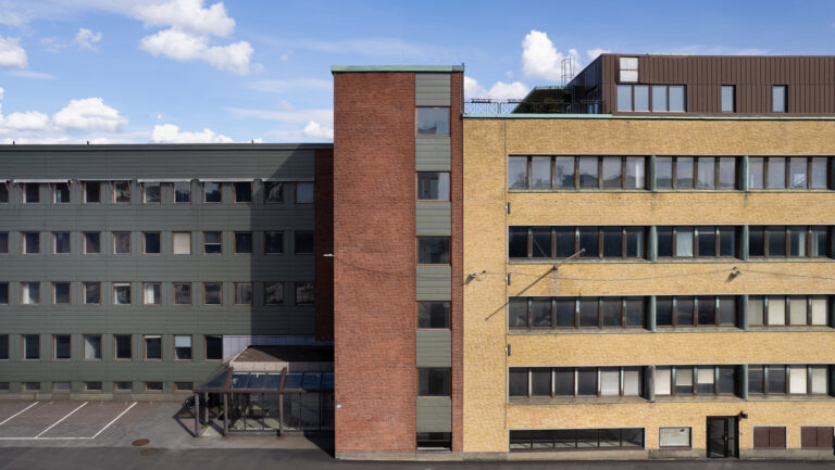 Fasad av en fler­våningsbyggnad med tre sammanhängande delar i grön, röd och gul tegel, med rader av fönster och blå himmel ovanför.