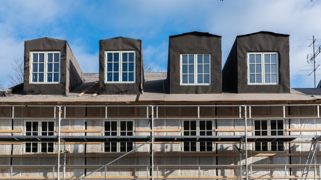 Fasad på ett flerbostadshus under renovering med byggställningar och fyra takkupor med fönster.