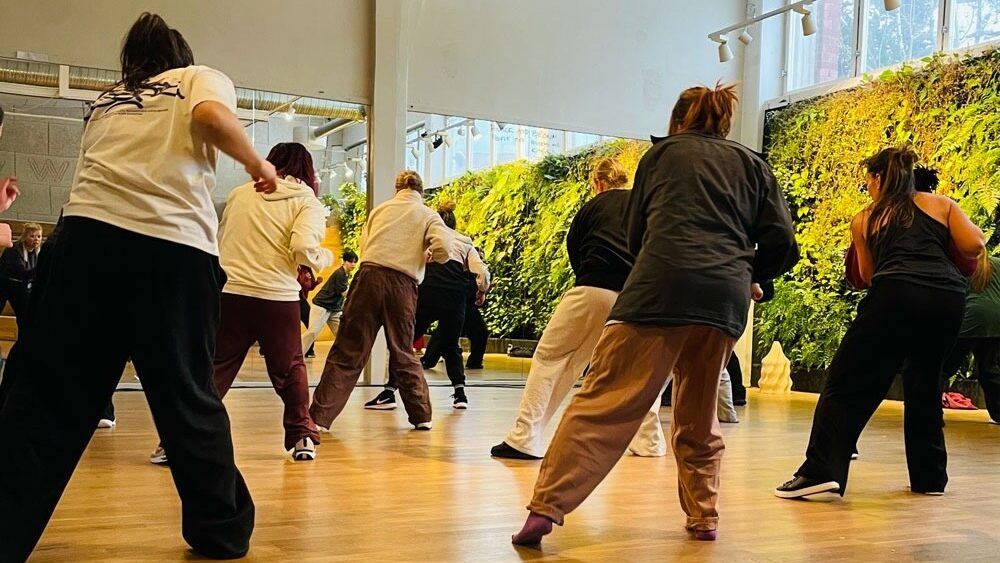 Ungdomar med säckiga kläder dansar med ryggarna mot kameran i en ljus danssal.