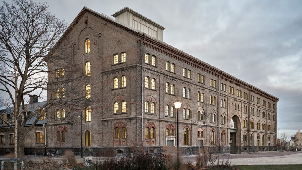 Tegelbyggnad i flera våningar med rundbågiga fönster och upplysta rum i skymningsljus, sedd snett från gaveln på byggnaden, med en stor portöppning och kala träd framför fasaden.