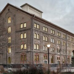 Tegelbyggnad i flera våningar med rundbågiga fönster och upplysta rum i skymningsljus, sedd snett från gaveln på byggnaden, med en stor portöppning och kala träd framför fasaden.