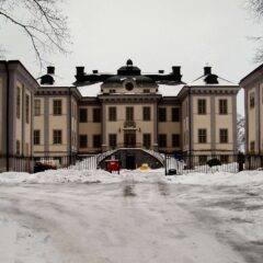 Snötäckt grusväg leder fram till en symmetrisk herrgårdsbyggnad med två flyglar, järngrind och barlösa träd under grå vinterhimmel.