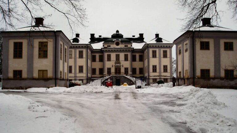Snötäckt grusväg leder fram till en symmetrisk herrgårdsbyggnad med två flyglar, järngrind och barlösa träd under grå vinterhimmel.