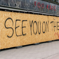 En person cyklar förbi ett högt träplank med där det står skrivet med svart See you on the other side