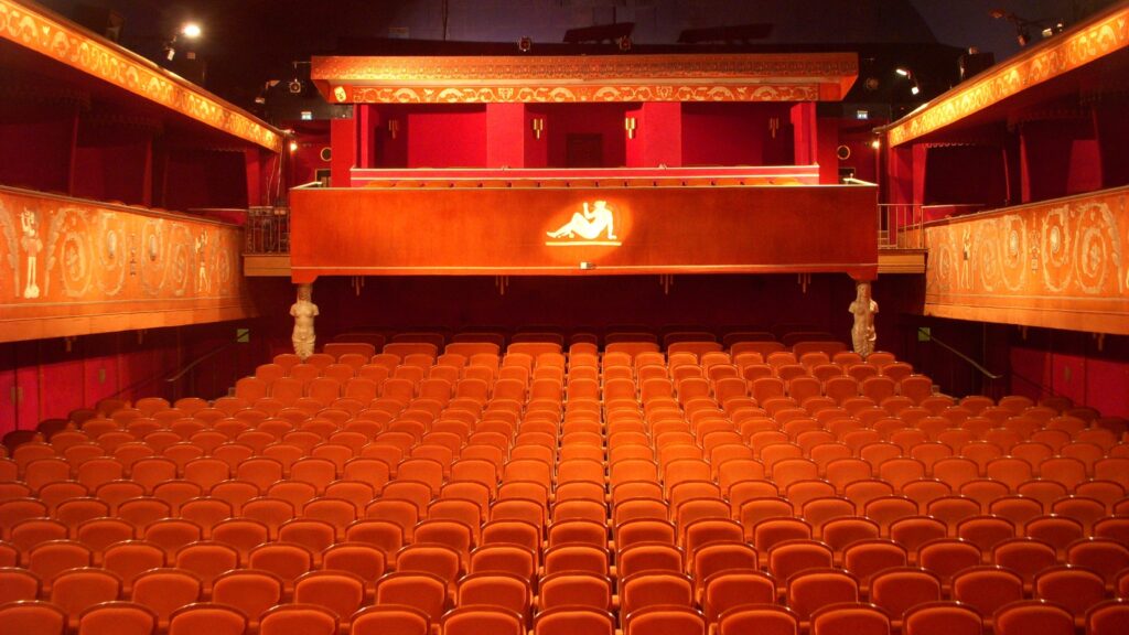 Interiör av en tom biografsalong sedd från scenen, med rader av orange-röda stoppade stolar som fyller parketten, två dekorerade balkonger längs sidorna och en bakre läktare. Väggar och balkongfronter är rikt utsmyckade i rött och guld, med varm belysning som lyser upp salongen.