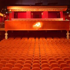 Interiör av en tom biografsalong sedd från scenen, med rader av orange-röda stoppade stolar som fyller parketten, två dekorerade balkonger längs sidorna och en bakre läktare. Väggar och balkongfronter är rikt utsmyckade i rött och guld, med varm belysning som lyser upp salongen.