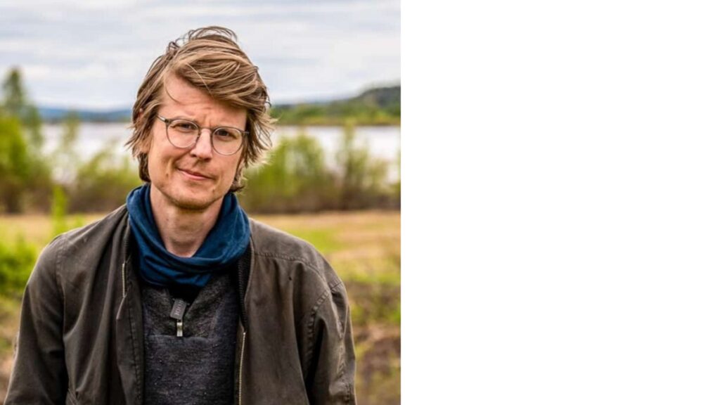 Porträttfoto på ung man med glasögon, i bakgrunden fält, skog och en älv.