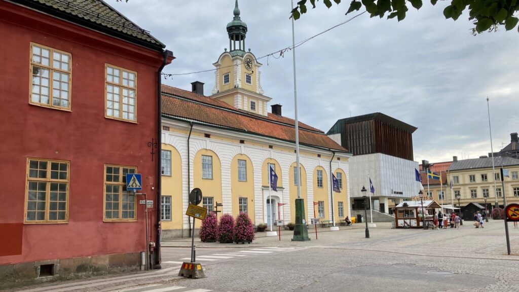 Ett stenlagt torg med en gul byggnad med klocktorn och flaggor, röda och gula hus runtom samt några människor och uteserveringar under molnig himmel.