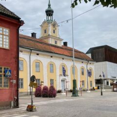 Ett stenlagt torg med en gul byggnad med klocktorn och flaggor, röda och gula hus runtom samt några människor och uteserveringar under molnig himmel.