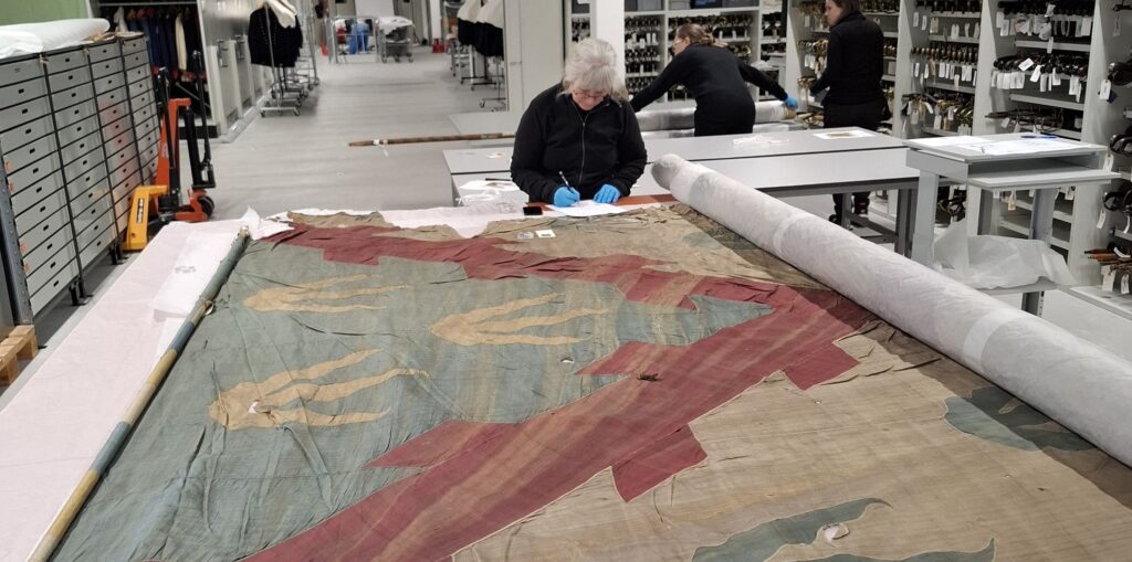 Ett slitet stort tygstycke ligger på ett vitt bord och en kvinna med blå plasthandskar undersöker