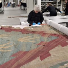 Ett slitet stort tygstycke ligger på ett vitt bord och en kvinna med blå plasthandskar undersöker