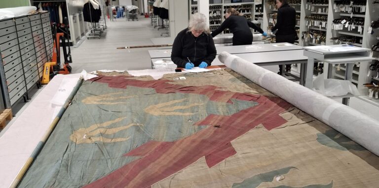 Ett slitet stort tygstycke ligger på ett vitt bord och en kvinna med blå plasthandskar undersöker