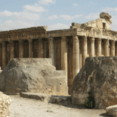 Antika stenruiner med höga kolonner i ett tempelområde, sedda från marknivå. I förgrunden ligger stora, väderbitna stenblock och i bakgrunden syns en lång rad kolonner mot en ljusblå himmel med några moln.