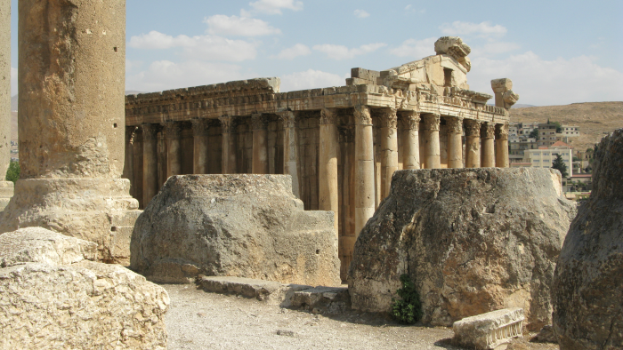 Antika stenruiner med höga kolonner i ett tempelområde, sedda från marknivå. I förgrunden ligger stora, väderbitna stenblock och i bakgrunden syns en lång rad kolonner mot en ljusblå himmel med några moln.