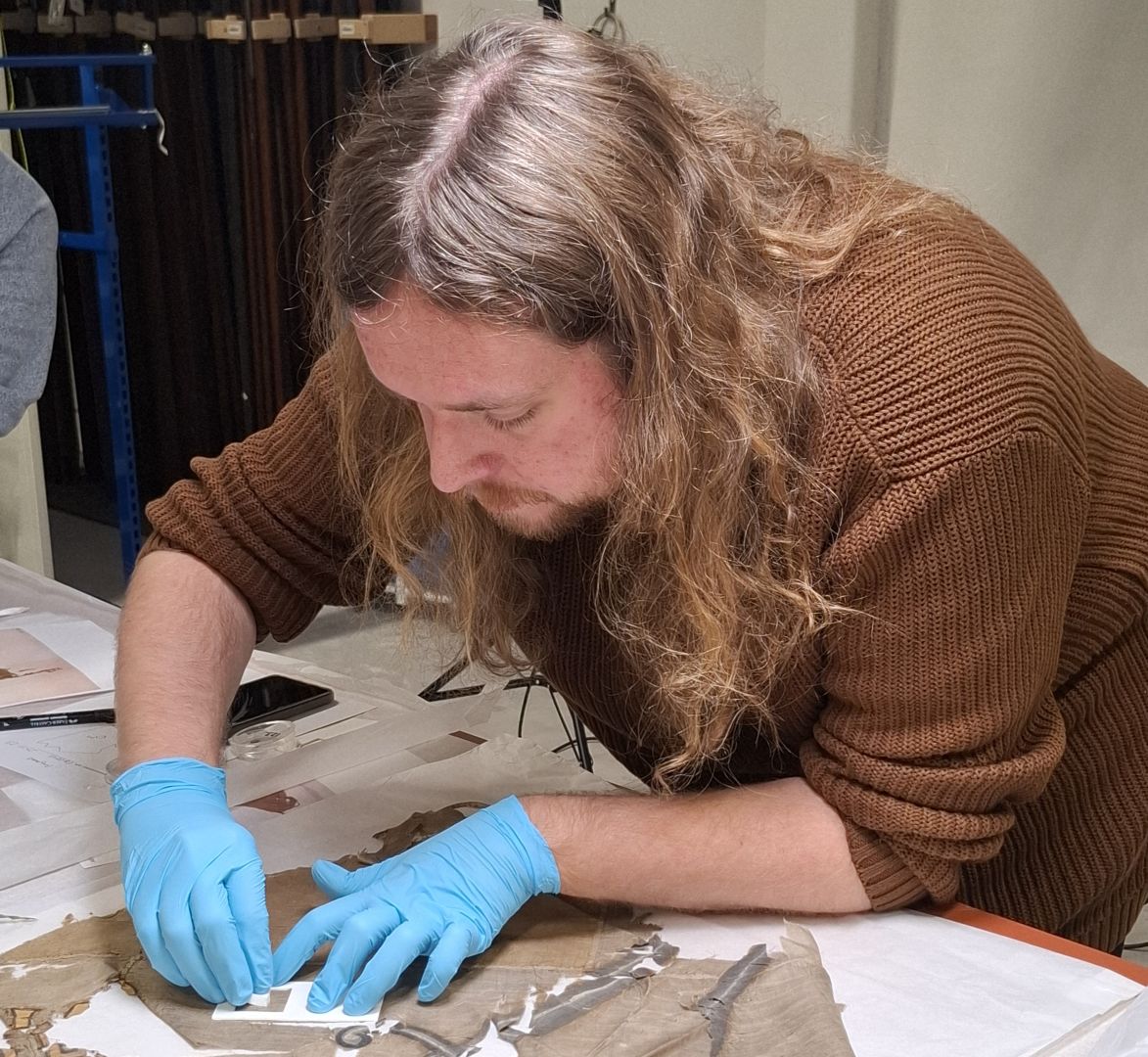 Ung man med långt brunt hår står böjd över ett slitet tygstycke och undersöker det med blå plasthandskar på sig