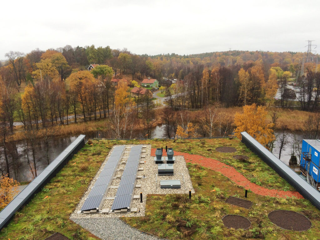 Vy över höstlandskap med färgade träd, små hus och en slingrande väg. I förgrunden syns ett tak med solpaneler och grön vegetation, nära en å.