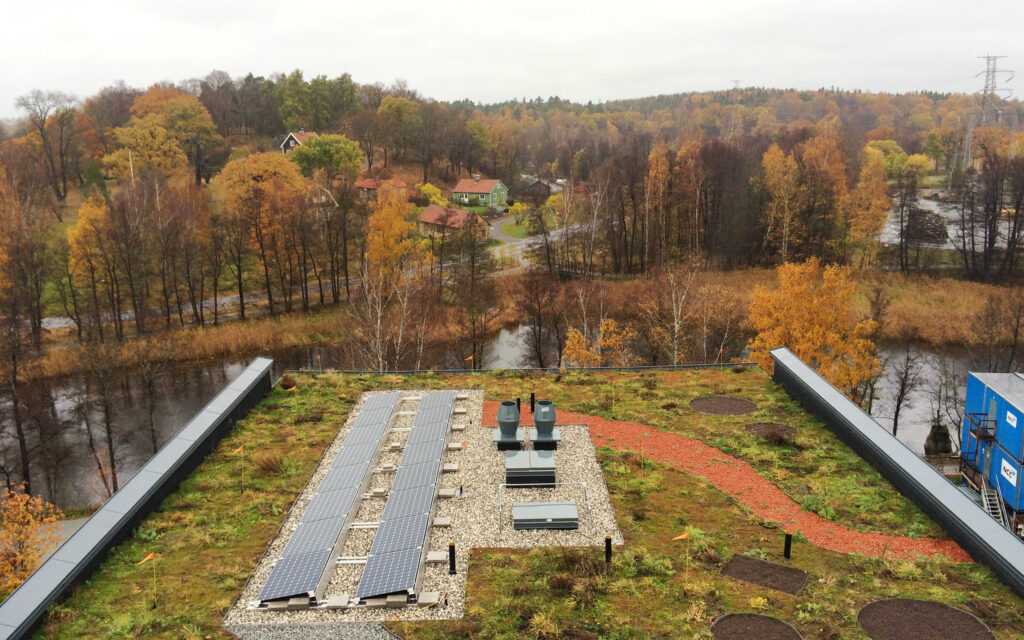 Vy över höstlandskap med färgade träd, små hus och en slingrande väg. I förgrunden syns ett tak med solpaneler och grön vegetation, nära en å.