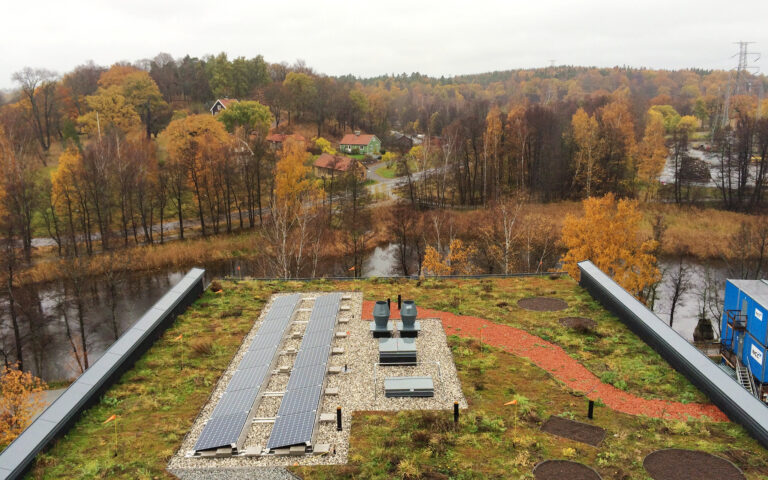 Vy över höstlandskap med färgade träd, små hus och en slingrande väg. I förgrunden syns ett tak med solpaneler och grön vegetation, nära en å.
