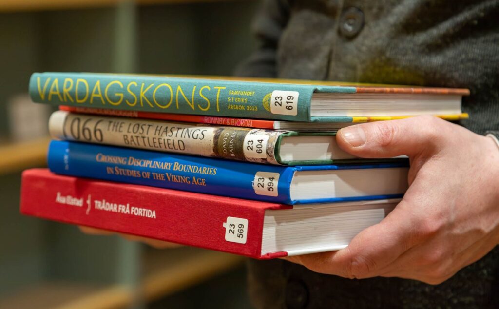 En bokhög där bokryggarna syns. Högen hålls av händer.