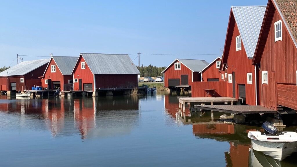Röda sjöbodar på pålar står tätt längs stilla vatten, med små bryggor och båtar intill, under klarblå himmel.