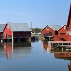 Röda sjöbodar på pålar står tätt längs stilla vatten, med små bryggor och båtar intill, under klarblå himmel.